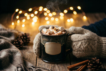 Hands in cozy knit mittens hold a mug of hot chocolate with marshmallows. A warm, festive winter scene with bokeh lights and cinnamon on a wooden table.