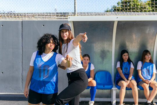 Soccer coach giving instructions to a female player on the field sidelines, the team listening from the bench