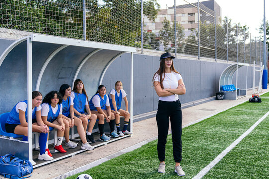 Female coach with crossed arms observing her women's soccer team sitting on the bench during a training session - Powered by Adobe