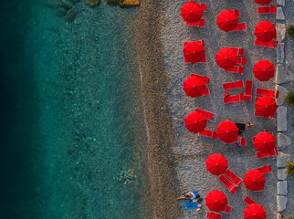 Aerial view of vibrant red umbrellas and beach chairs pop against the turquoise sea and pebbled shore, a summer haven of color and relaxation,Sistiana, Friuli-Venezia Giulia, Italy.