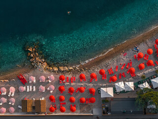 Aerial view of vibrant red umbrellas line a sun-drenched beach, contrasting with the turquoise sea and rocky shore, framed by verdant trees, Sistiana, Friuli-Venezia Giulia, Italy.