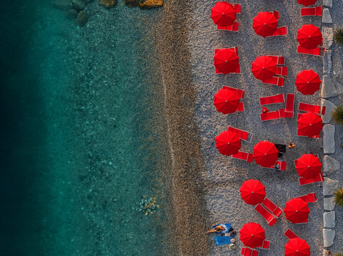 Aerial view of vibrant red umbrellas and beach chairs pop against the turquoise sea and pebbled shore, a summer haven of color and relaxation,Sistiana, Friuli-Venezia Giulia, Italy.