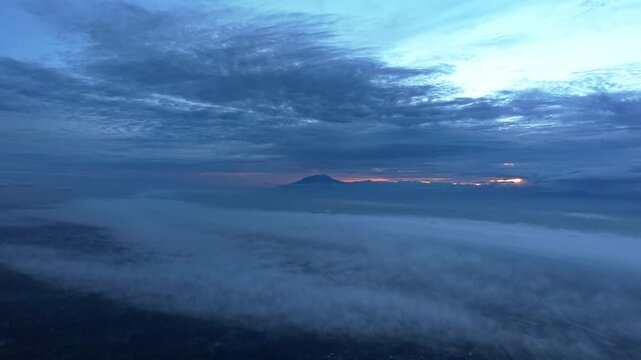 Aerial view of a solitary mountain peak piercing through a sea of fluffy white clouds under a pastel sky, Klaten Regency, Central Java, indonesia.