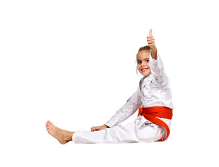 A karate girl sits on the floor in a kimono with an orange belt and shows a thumbs up