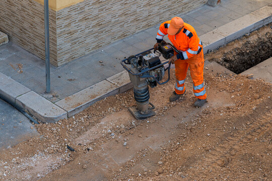 Top view of a construction worker in orange safety gear using a tamping rammer to compact gravel in a trench next to a sidewalk during urban infrastructure repairs.