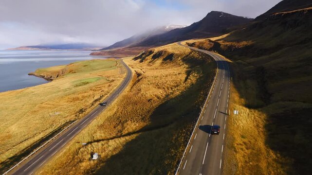 Vehicles driving at sunset along a cliffside road in the fjords, with the ocean beside it. Scenic landscape with warm colors and dramatic coastal views.