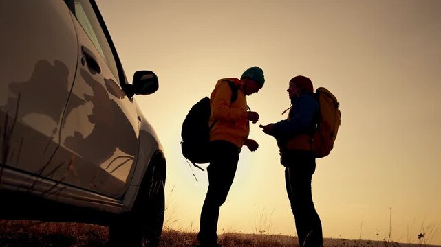 Hikers talk near parked car during sunset, two backpacks and silhouette on roadside, couple sharing conversation and checking map, travel and road pause in open nature, hiking couple return car at