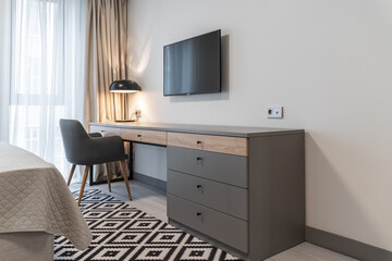 next to bed in bedroom is gray table with drawers and chair. A desk lamp is on on the table, and a TV is on the white wall. There is a white carpet with a pattern of black rhombus on the floor.