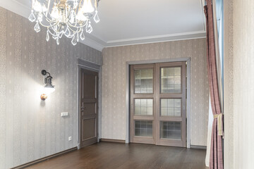 elegant empty room with wooden doors, wooden brown floors, wallpaper and retro lamps on the wall, and a chandelier with glass pendants on the ceiling. The room has a cozy dim light from the lamps.