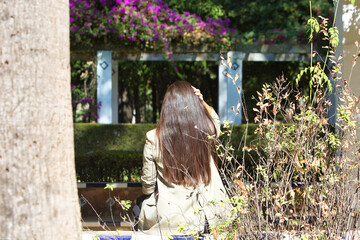 Photo taken from behind of a woman sitting on a bench among the vegetation. The woman with long, dark hair is touching her hair with her hands.