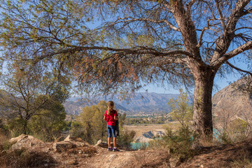 Young girl in red t-shirt framed by forest branches, looking at reservoir water, childhood discovery and nature exploration
