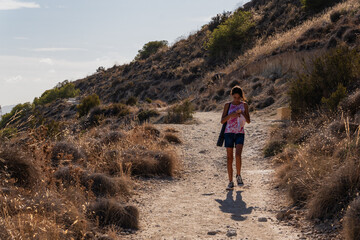 Young girl walking on a natural hiking trail with rock pavements while looking at her mobile phone. Technology and nature contrast.