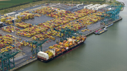 Aerial view of a massive cargo ship docked beside a vibrant port, filled with stacks of colorful containers under the watchful gaze of towering cranes, Antwerp, Vlaanderen, Belgium.