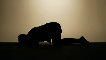 Kneeling silhouette lowering forehead toward mat in studio, barefoot wearing shorts backlit halo