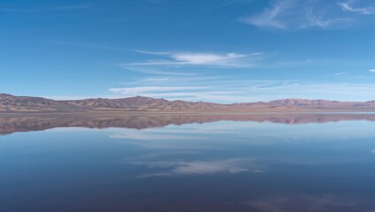 Fototapeta premium Reflecting calm lake surface mirroring low mountain ridge at remote basin, with wispy cirrus clouds
