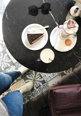 Cozy Coffee Break with Cake and Accessories. Top-down photo of a cozy café setting featuring a marble table with a slice of chocolate cake, a cappuccino with whipped cream and cocoa