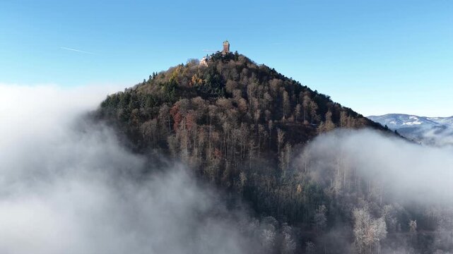 Aerial view of Chateau de Ramstein atop a forested peak, veiled in ethereal mist, with distant mountains under a clear sky, Orschwiller, Grand Est, France.