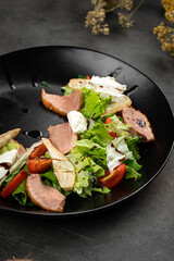 Fresh duck salad with pear slices, tomatoes, leafy greens and creamy cheese on a dark plate