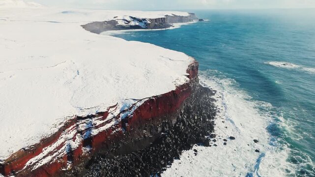 Aerial view of storm ocean waves crashing against snowy coastal cliffs at winter sunrise. The Icelandic plateau reveals striking, martial red mineral cliffs contrasted against the icy landscape