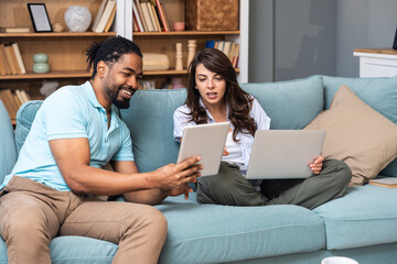 Smiling couple sits on a sofa comparing a tablet and laptop, coordinating tasks for budgeting,...