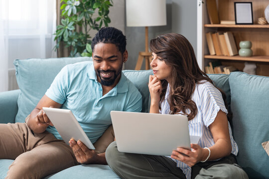 Smiling couple sits on a sofa comparing a tablet and laptop, coordinating tasks for budgeting, study, or a small business project. Real home collaboration, remote work energy, positive teamwork.
