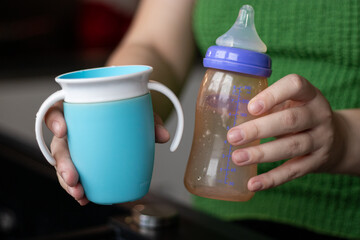 Close up of female hands holding a bottle and a no spin cup for baby milk 