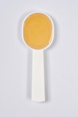 A white plastic measuring spoon holds a dose of golden-yellow cough syrup or liquid medicine, photographed from above on a light gray background