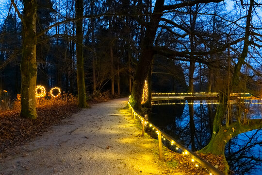 Magical forest path illuminated by warm yellow Christmas lights along the railings and trees, reflected in a calm pond at dusk. Atmospheric winter evening scene