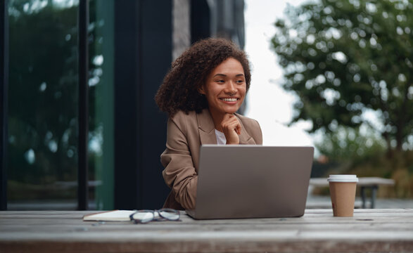black recruiter smiling during video call with laptop on outdoor table, coffee cup beside device, attentive