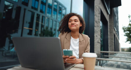 young black woman with laptop smiling while checking smartphone, outdoor cafe setting, coffee cup, modern