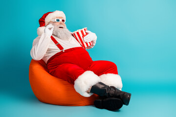 Santa relaxes in a bright blue studio holding popcorn wearing festive 3d glasses on an orange beanbag for christmas promo