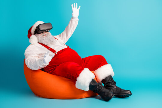 Santa Claus in a virtual reality headset relaxes on an orange bean bag against a bright blue background creating a festive holiday moment of fun and imagination