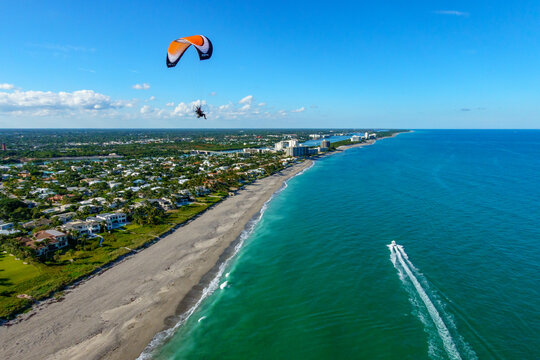 Aerial view of a paraglider soaring over the turquoise ocean as a boat speeds along the coast near the buildings, Jupiter, Florida, United States.