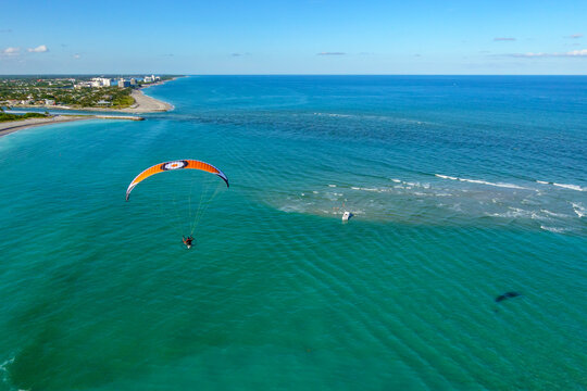 Aerial view of a paraglider soaring over the turquoise water where the ocean meets the land, contrasting with the beach and buildings, Jupiter, Florida, United States.