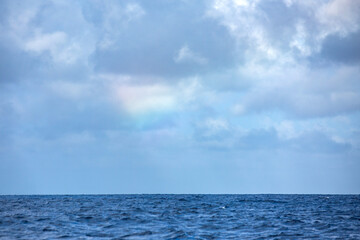 An idyllic tropical blue ocean featuring a feint rainbow in the storm clouds above the horizon.
