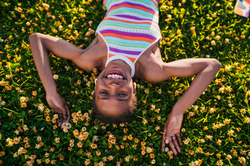 African woman laying on her back in blooming grass, smiling brightly at camera with smartphone...