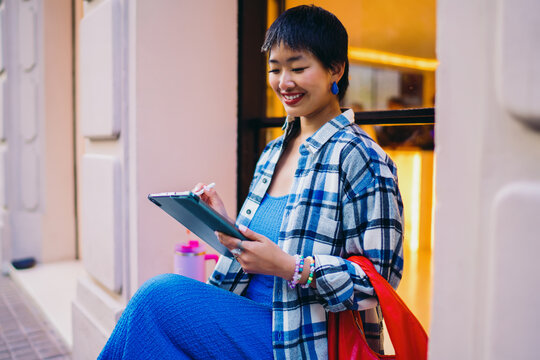 Asian woman seated on urban bench smiling gently while using stylus on digital tablet, wearing plaid and blue, representing peaceful digital engagement and self-expression.