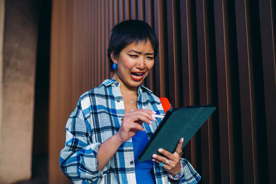 Surprised Asian woman reacting emotionally to digital content, holding stylus and tablet, capturing real-time feedback in dynamic mobile workspace setting.