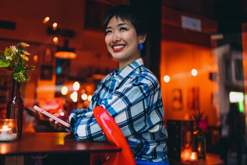 Smiling woman in plaid shirt leans on table with smartphone, embracing warmth and creativity in digital space, surrounded by café lights and feminine freelance energy.