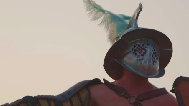A cinematic, dramatic close-up of a Roman gladiator in detailed armor and crested helmet (galea), slowly raising a curved sword (sica) with a focused, intense action shot against a hazy sky.