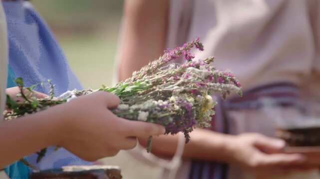 Close-up shot of two people's hands: one holding a small, steaming clay bowl (incense, smudging), and the other holding a freshly braided wreath of dried wild herbs and flowers.