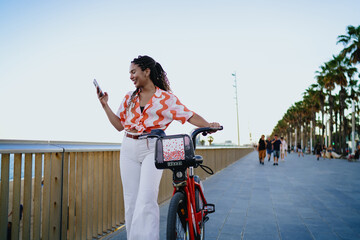 Smiling young woman with braids holding smartphone while resting on red rental bike, symbolizing digital lifestyle, modern mobility and joyful independence in everyday technology use.