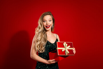 Fototapeta premium Young woman in green sequin dress presents festive red gift box with gold ribbon against red background