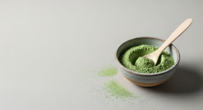 A bowl of vibrant green matcha powder sits beside a wooden spoon on a clean gray surface perfect for food blogs or wellness publications
