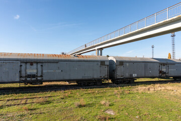 Fototapeta premium Rusty freight train cars parked on grassy tracks beneath a modern pedestrian bridge against a clear blue sky.
