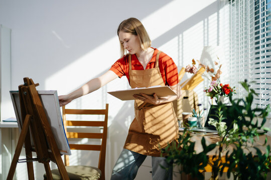 A female artist painting on canvas in a bright home studio, expressing creativity and focus.