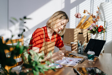 A focused female artist painting at a wooden desk in a bright home studio with art tools, laptop, and natural sunlight
