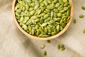 Raw peeled green pumpkin seeds in wooden bowl on natural linen fabric background top view. Food background, organic healthy tasty pumpkin seeds, autumn cooking ingredient