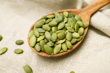 Raw peeled green pumpkin seeds in wooden spoon on natural linen fabric background top view. Food background, Organic healthy tasty pumpkin seeds, Autumn cooking ingredient. Plant-based protein source