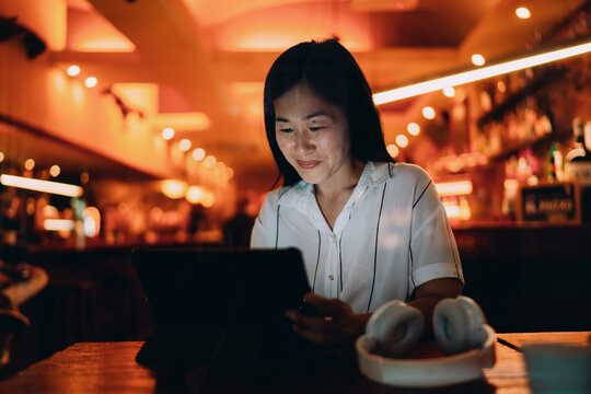 Focused Asian woman using tablet in warm-lit cafe, reflecting freelance lifestyle, digital workspace and creative productivity in modern tech environment.